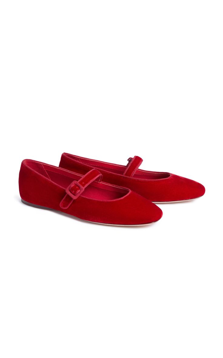 a pair of red velvet mary jane flats are displayed on a white background. the shoes have a pointed toe and a small buckle strap across the instep. they are positioned slightly apart, with one shoe slightly in front of the other.  the shoes appear to be made of a soft, luxurious velvet material.