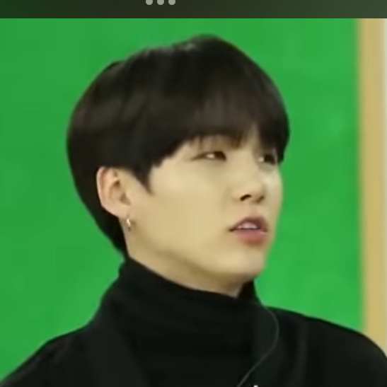 a headshot of a young man with black hair wearing a black turtleneck sweater and a small earring. he is looking slightly to the right and has a neutral expression. the background is green.