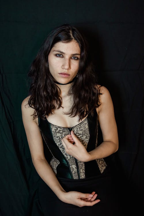 closeup portrait of a young woman with dark, wavy hair wearing a dark green and beige lace corset. she is sitting against a dark background, her expression is serious, and she is looking directly at the camera. her hands are gently touching the front of her corset. the overall mood is mysterious and elegant.