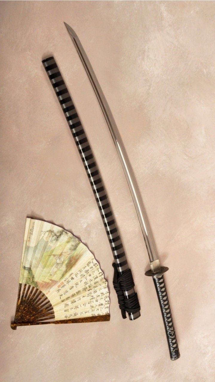 a katana and a japanese fan are displayed on a light pink surface. the katana is out of its sheath, lying next to it. the sheath and handle are black and silver. the blade is shiny and silver. the fan has a painted scene and writing on it.