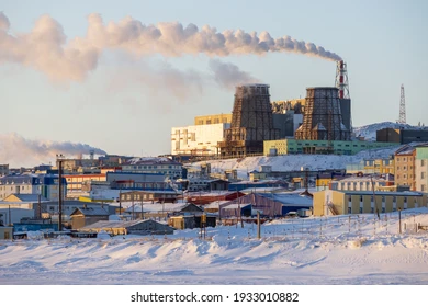 a panoramic view of a city in the arctic, dominated by a large industrial plant. the factory emits a plume of smoke into the clear blue sky, with a few puffy clouds hanging in the air. the buildings are covered in snow, and the ground is white. there is a sense of cold and isolation, as the city appears to be located in a remote, desolate environment.