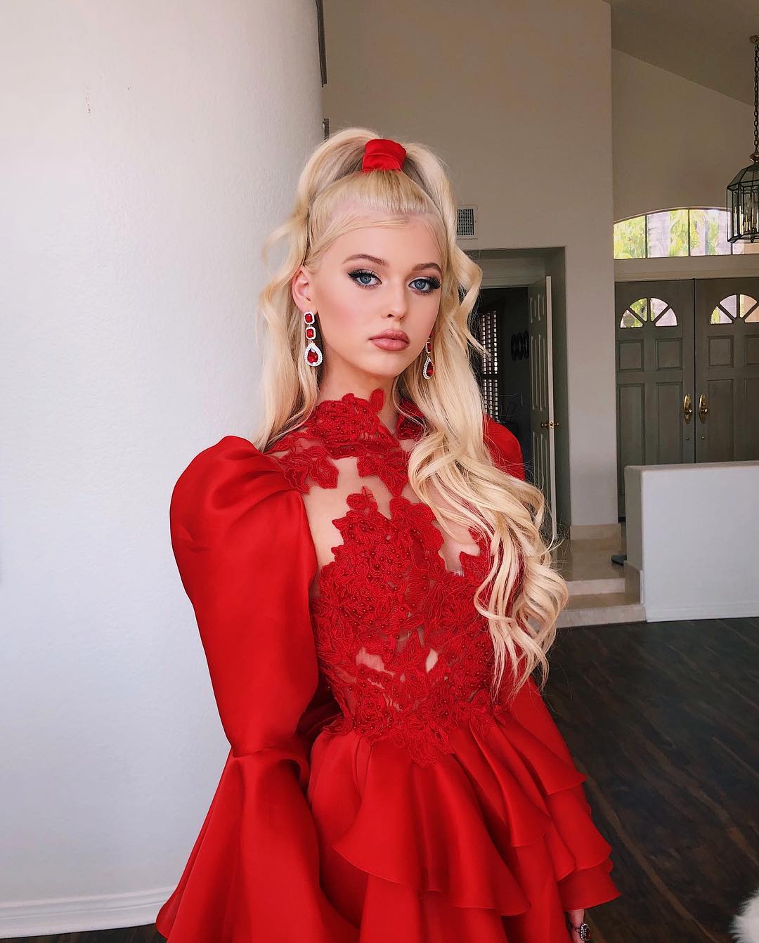 a woman with blonde hair in a ponytail wearing a red dress and earrings is posing in front of a house interior. she is facing the camera. the dress has lace detailing and puffy sleeves. there is a door and chandelier in the background.