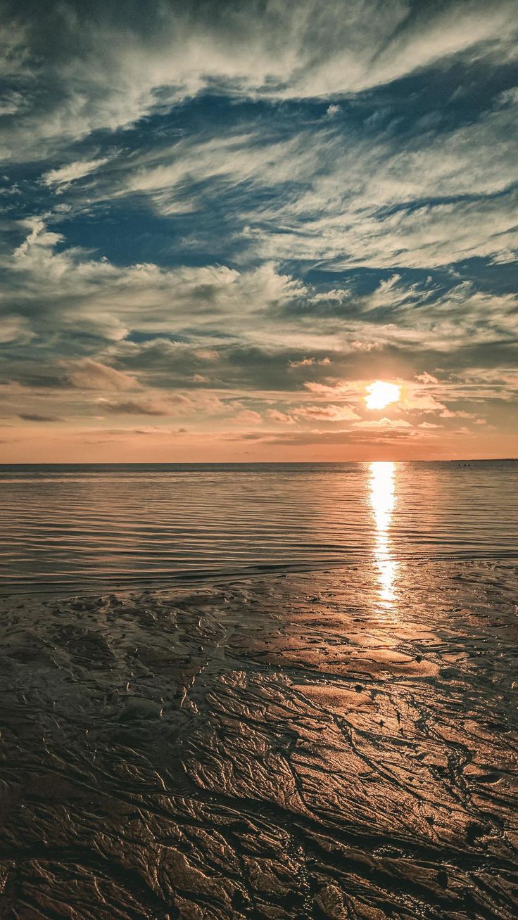 a scenic shot capturing a tranquil beach at sunset. the golden sun reflects brightly on the calm water, creating a shimmering effect. the sky is filled with a mix of white and gray clouds, tinted with orange hues from the setting sun. the foreground shows textured sand, wet from the receding tide. the overall scene evokes a sense of peace and natural beauty.
