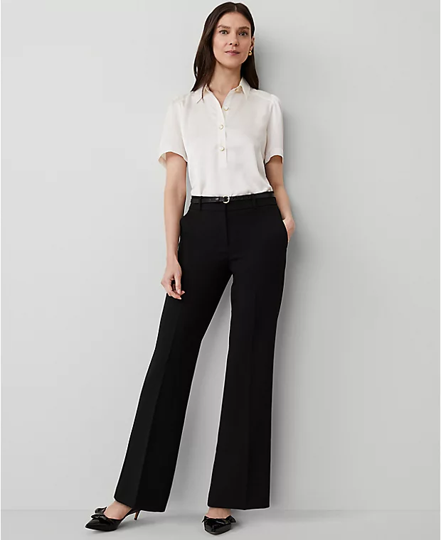 a woman in a white shirt and black pants is standing in a studio with her arms at her side. she is wearing a black belt and black shoes. she is looking at the camera.