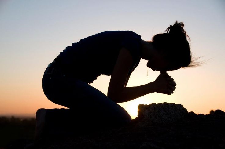 a silhouette of a woman kneeling on a rock, her head bowed in prayer, as the sun sets in the background.