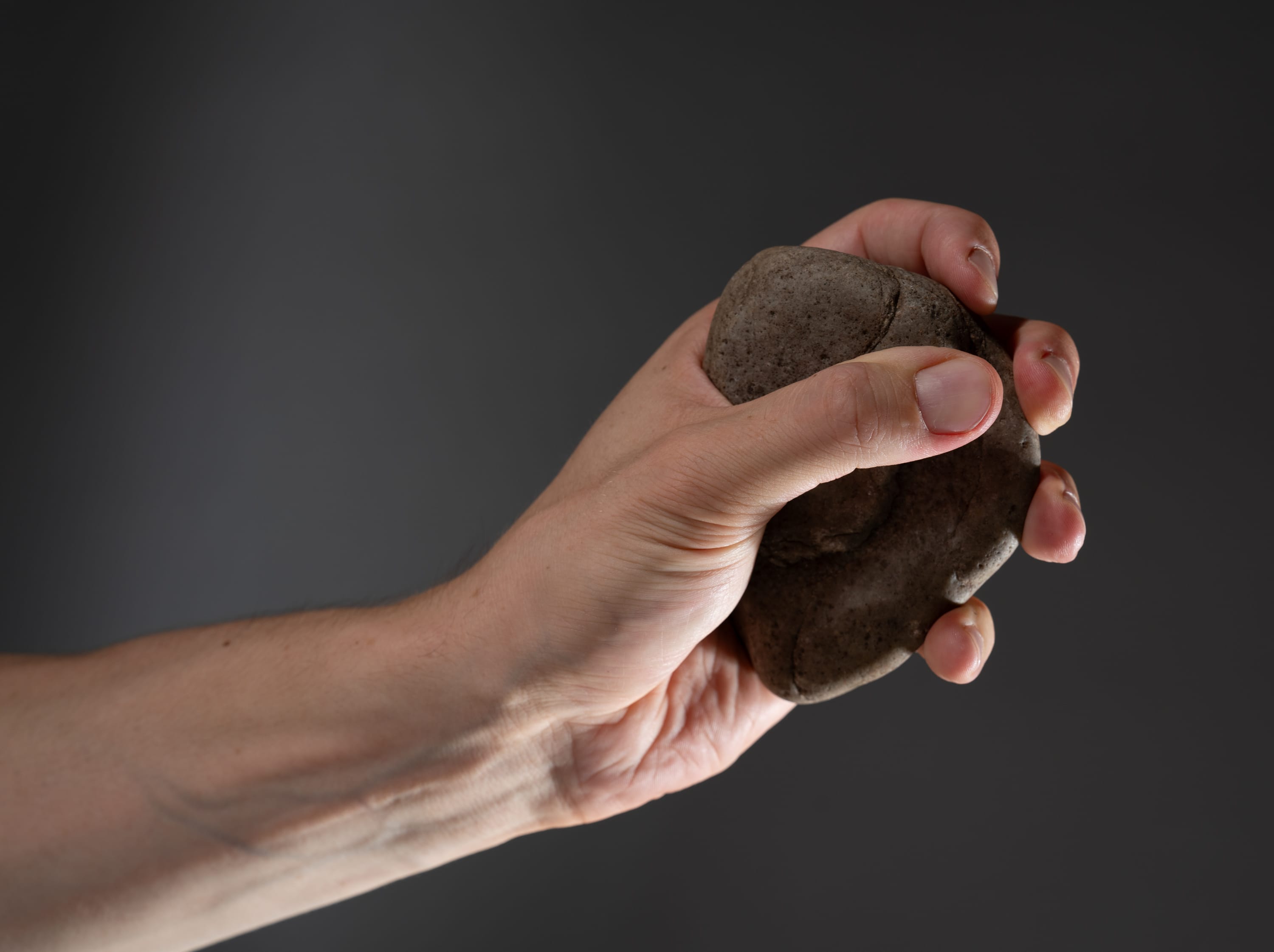 a closeup shot of a hand tightly gripping a rough, gray rock against a dark gray background. the studio lighting highlights the texture of both the rock and the hand.