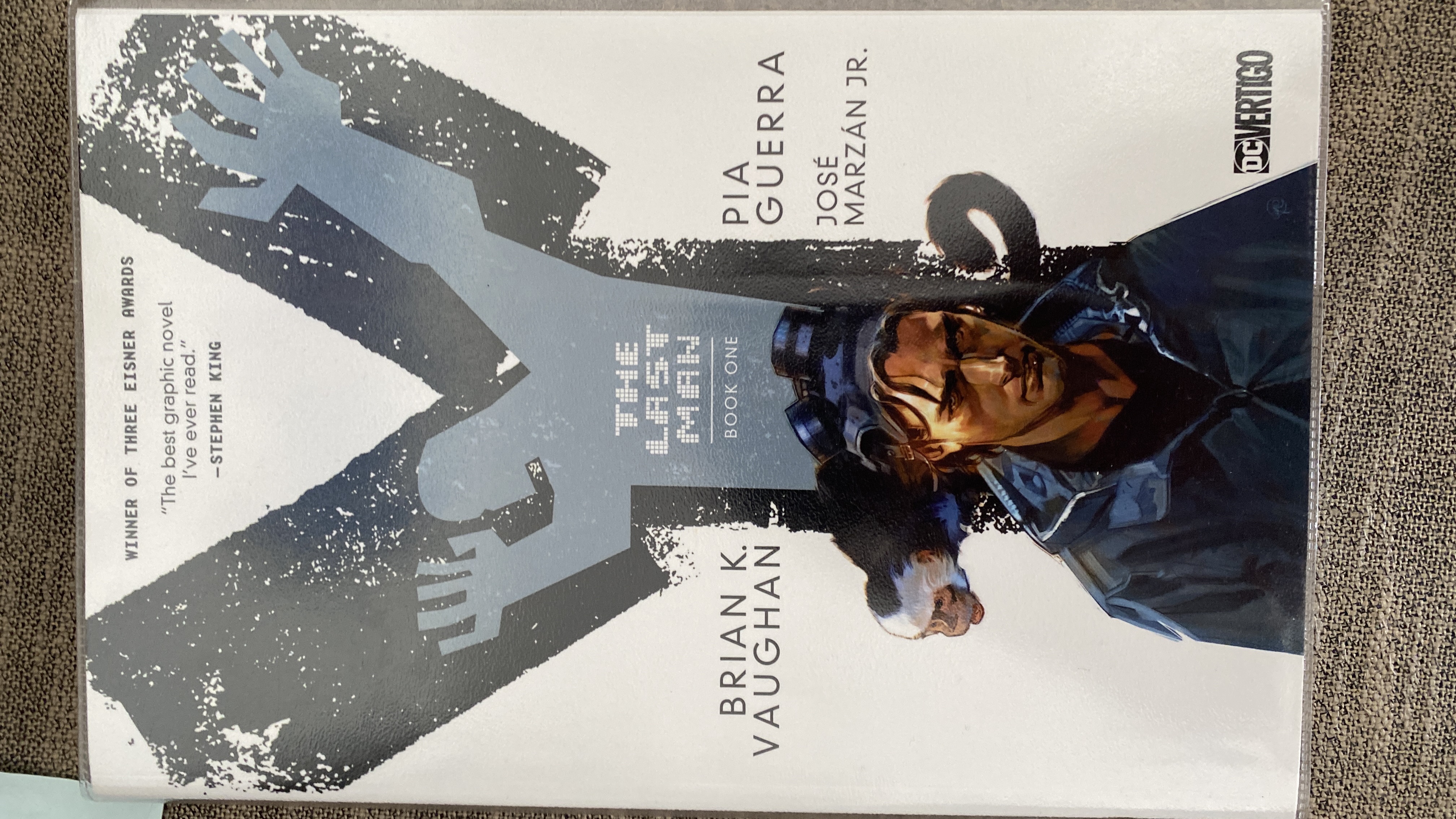 the image is the cover of the graphic novel "the last man" by brian k. vaughan and pia guerra. it features a man standing in a futuristic setting, with a shadowed figure in the background and a monkey in the foreground. the cover also includes the title of the book, the authors' names, and the publisher's logo. the book is set in a dystopian future where a plague has wiped out almost all of humanity. the main character is a man who is trying to survive in this new world and find out what happened to the rest of the world. the story is full of action, adventure, and suspense.