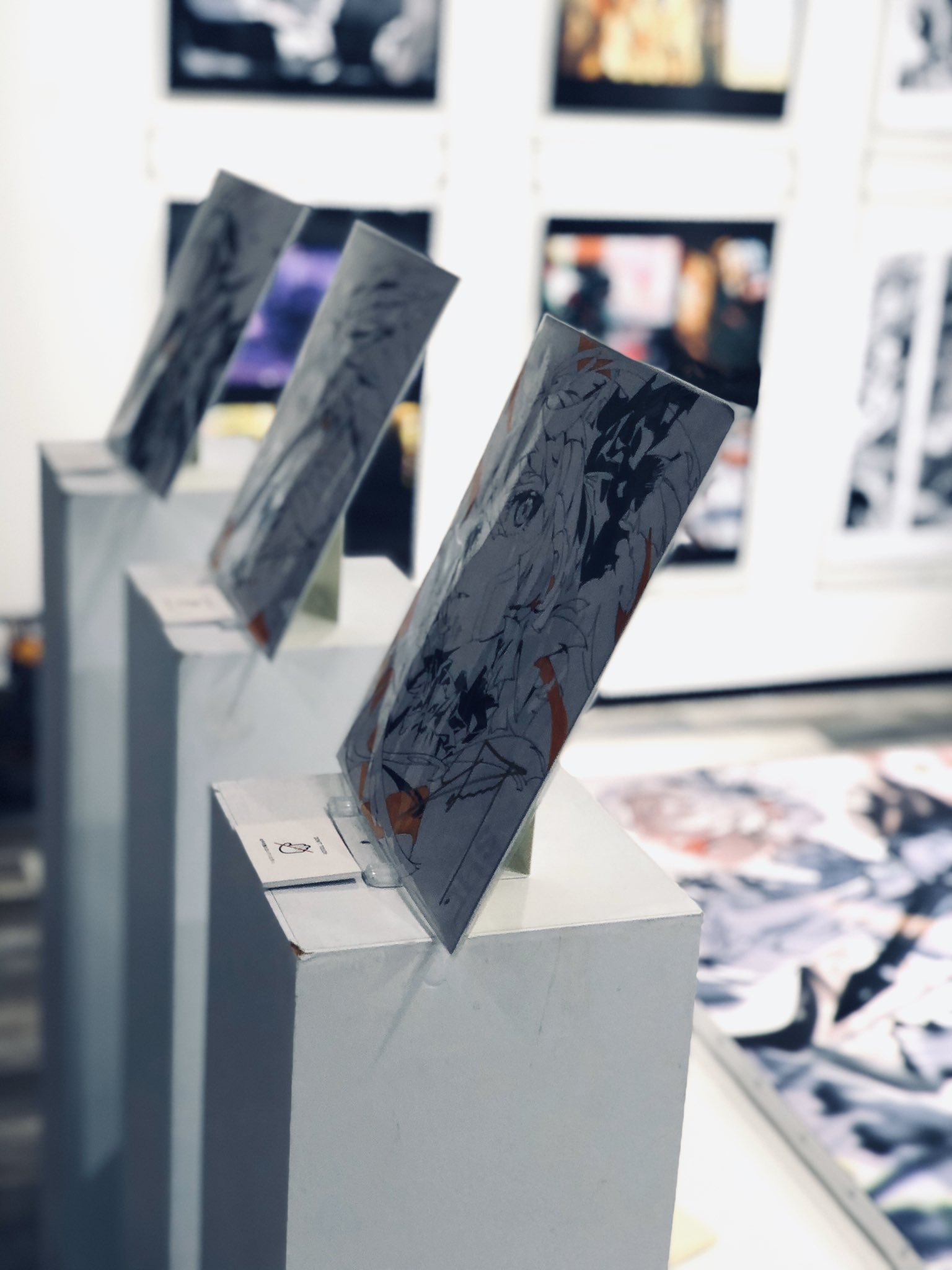 the image shows a closeup view of several framed illustrations displayed on white pedestals in an art gallery setting. the illustrations are in black and white and appear to be in an anime or manga style. the focus is on the texture and details of the artwork, with a shallow depth of field blurring the background. the overall ambiance is modern and minimalist, highlighting the artwork.