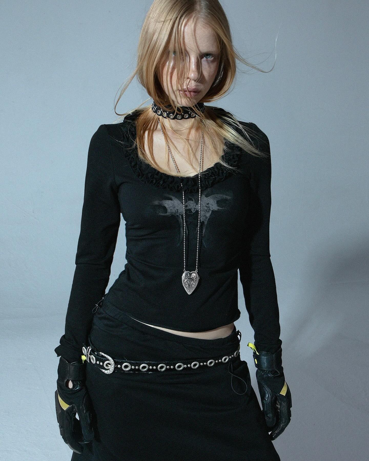 the image shows a young woman with long blonde hair wearing a predominantly black outfit.  her style is gothic or alternative, featuring a longsleeved black top with a subtle design, a black skirt, a studded silver belt, a choker necklace, a silver heart pendant necklace, and black gloves with yellow accents.  she is posing against a plain light grey background, giving a serious expression to the camera. the overall aesthetic is dark, stylish, and edgy.