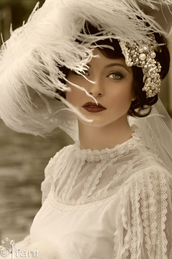 a beautiful woman in a vintage white dress and a hat with feathers. she is looking at the camera with a soft, dreamy expression. the dress is made of delicate lace and the hat is adorned with feathers and pearls. she is wearing a beautiful diamond and pearl jewelry. she is very glamorous and elegant. the woman has beautiful make up, with a soft pink blush and a red lipstick. she has curled her hair and is wearing a headband.