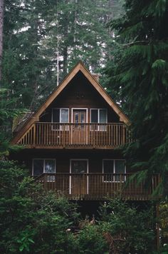 a twostory wooden aframe cabin nestled in a dense forest. the cabin features a porch/balcony on both levels, with windows and a door on the top floor. the surrounding woods are lush and green, creating a secluded and rustic atmosphere.