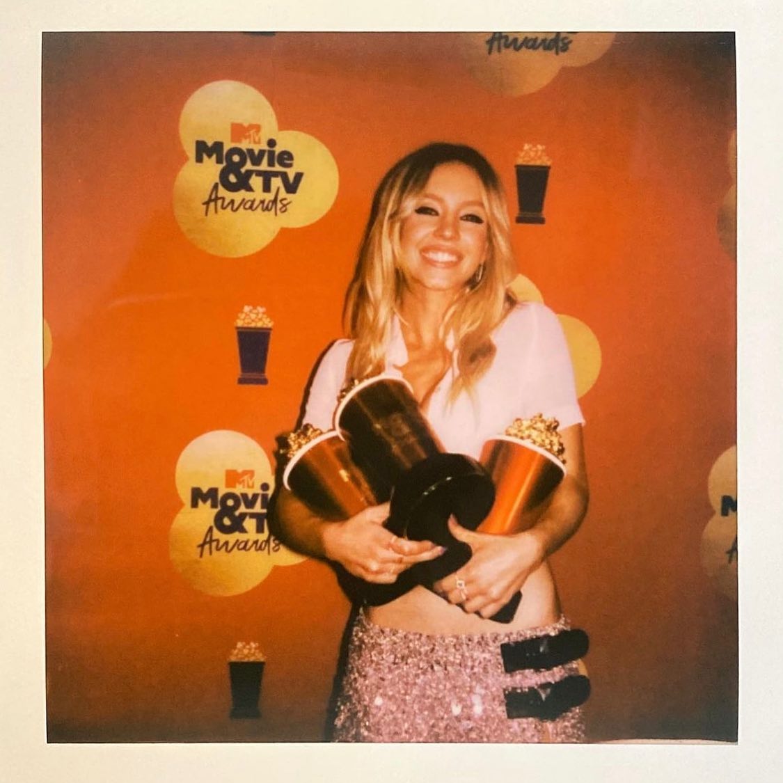 the image is a polaroidstyle photograph of a blonde woman at the mtv movie & tv awards. she is smiling and holding several popcornshaped awards. she's wearing a light pink shirt and a sparkly skirt.  the background is an orange backdrop typical of award show events, with the mtv movie & tv awards logo visible. the overall feel is celebratory and joyful.