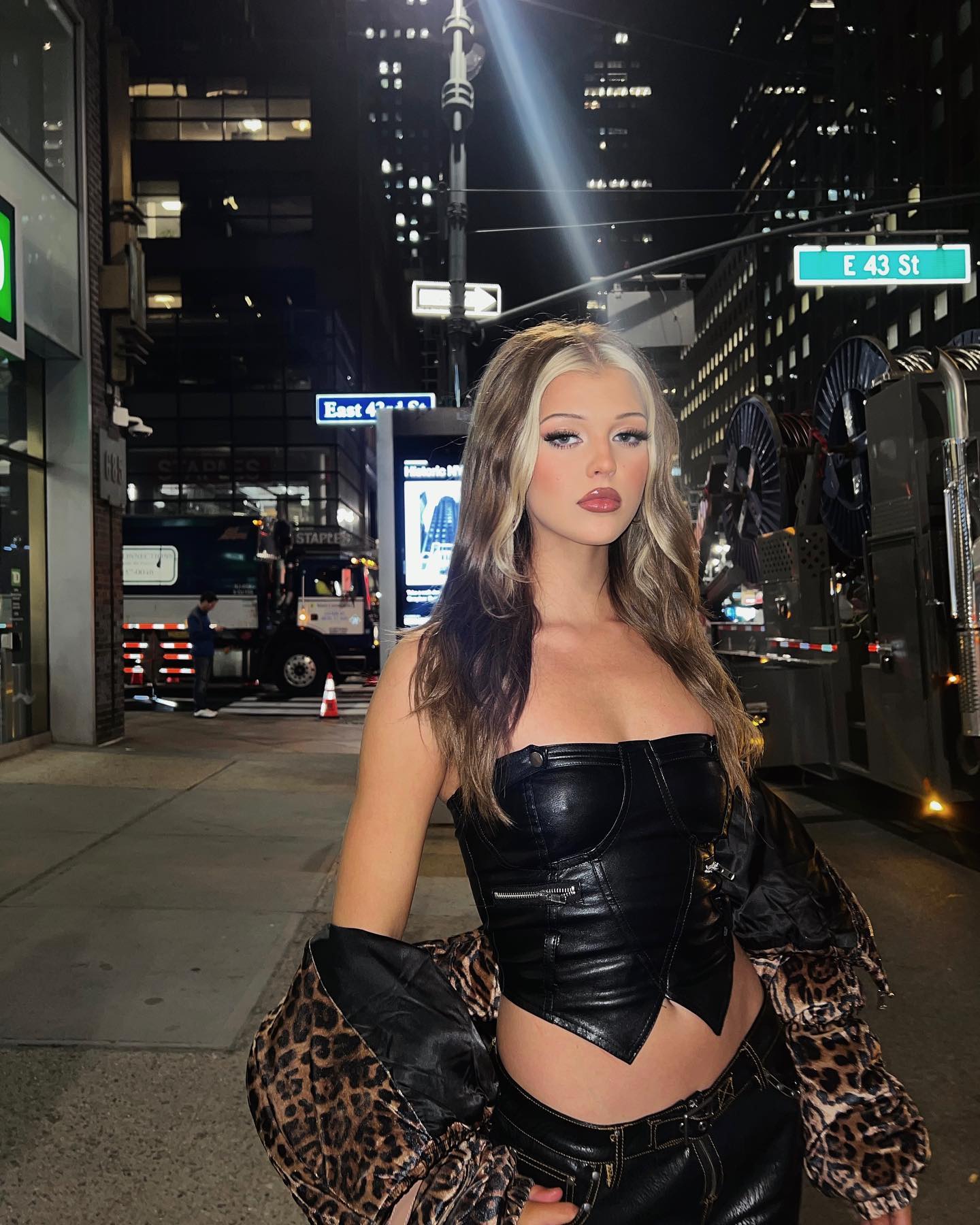 a young woman with blonde highlights in her hair is standing on a city street at night. she is wearing a black corset top, black pants, and a leopard print jacket. behind her are buildings, a street light, and a truck. a street sign reads "east 43 st."