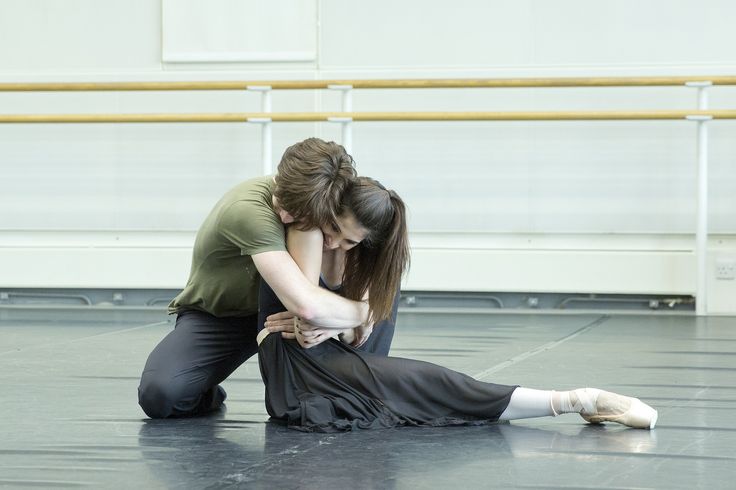 a ballet dancer in a black tutu is being comforted by her male partner in a green shirt. the dancer is sitting on the floor with her leg extended while the male partner is kneeling in front of her with his arms wrapped around her. they both have their heads bowed in a pose of sadness. they are in a dance studio setting with a wooden barre and a white wall in the background.