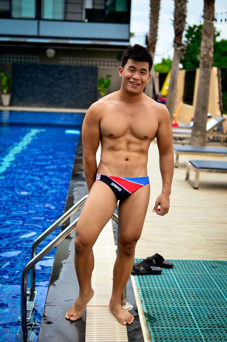 a young, muscular man in a pair of arena swim trunks stands by the edge of a swimming pool. he is smiling and appears to be enjoying himself. the setting is a resort or hotel with palm trees and loungers visible in the background.  the man is wellbuilt and has a healthy tan.  the overall mood of the image is relaxed and carefree.