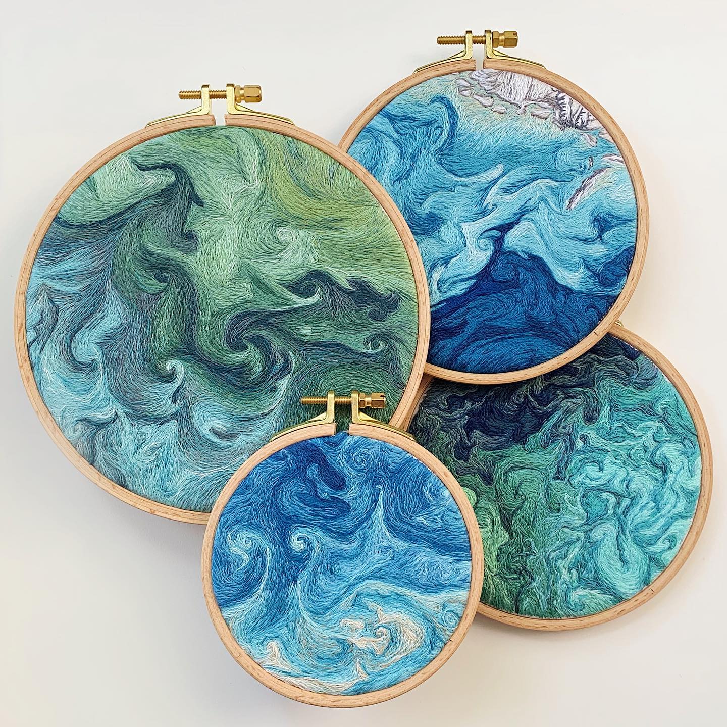 the image showcases four embroidery hoops of varying sizes, each displaying a mesmerizing abstract representation of ocean waves. the skillful use of blue, green, and turquoise threads creates a sense of depth and movement, mimicking the fluidity and power of the sea. the hoops, crafted from lightcolored wood, provide a natural frame for the intricate artwork, enhancing the overall visual appeal. the arrangement of the hoops, with their overlapping compositions, adds a dynamic and captivating element to the display.  the image is bathed in soft, natural light, accentuating the textures and subtle color variations within the embroidered artwork.