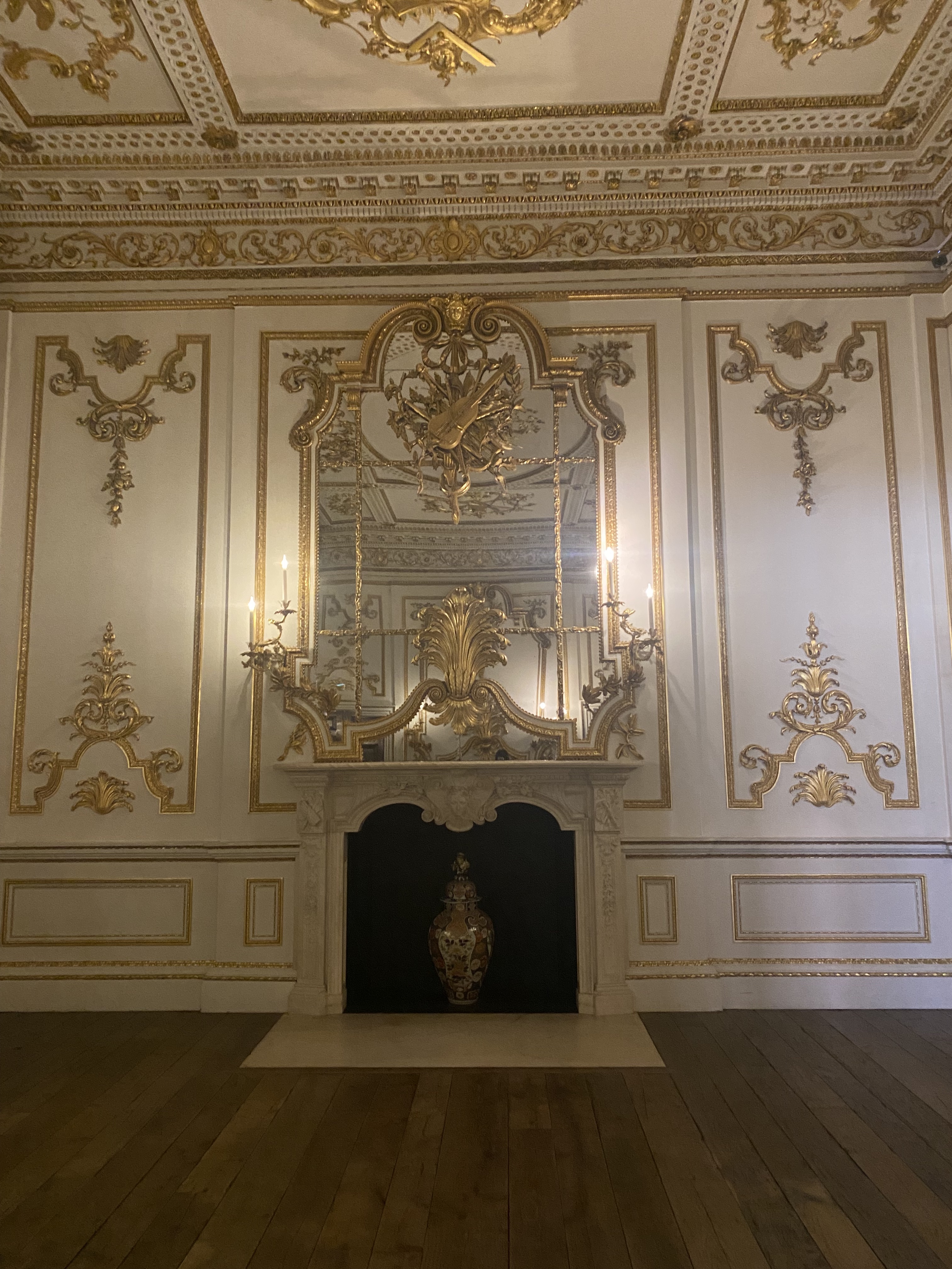 the image shows an ornate room with a rococo style design. the walls are white with gold trim and detailing. a large fireplace with a mirror above it is the focal point of the room. a vase sits inside the fireplace. the ceiling is also highly decorated with gold details. the floor is made of hardwood.