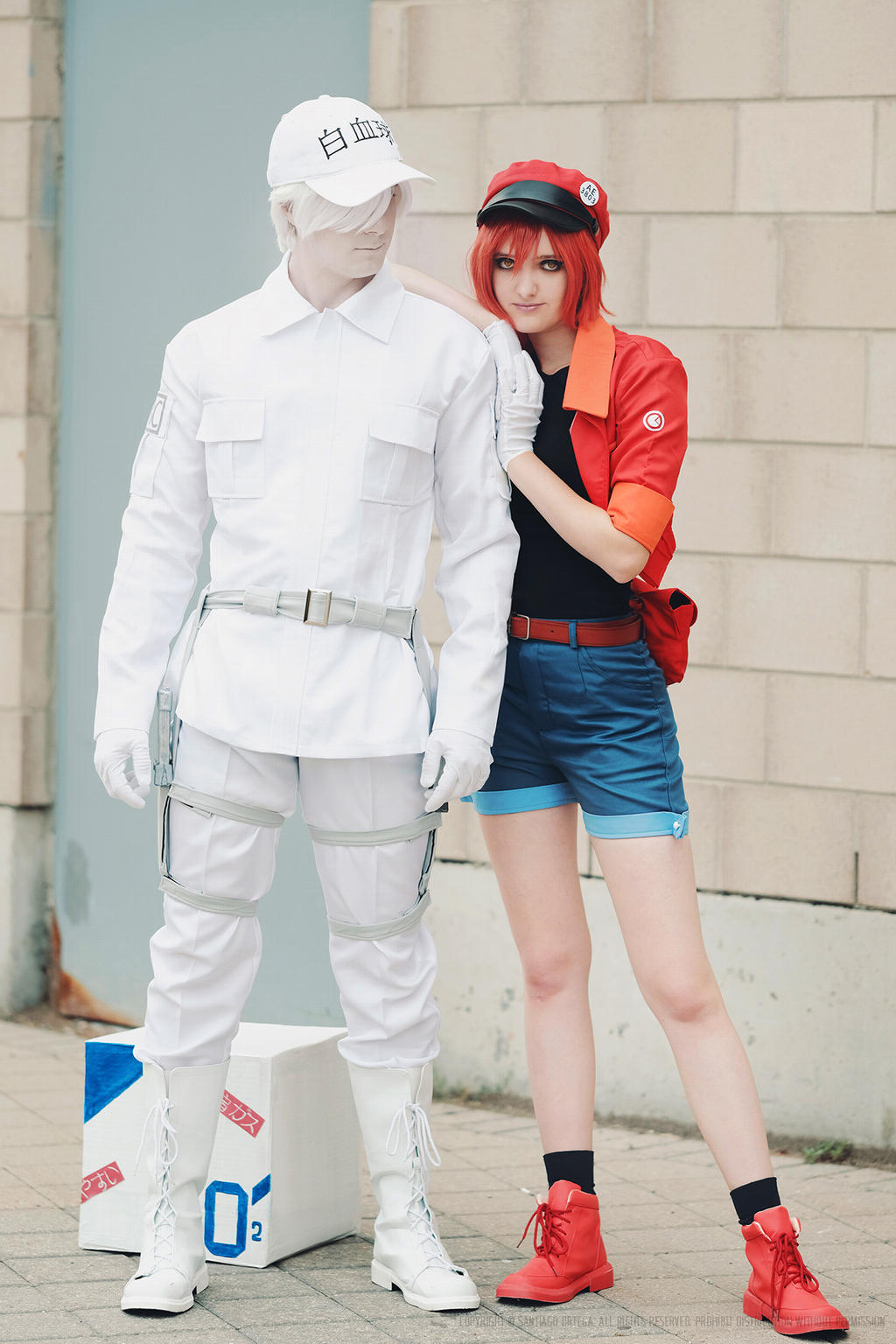 a full shot captures two cosplayers in character from the anime "cells at work!". on the left stands a person dressed as white blood cell, sporting a complete white outfit consisting of a longsleeved shirt, pants, gloves, boots, and a cap with japanese writing. straps and holsters add to the outfit's detail. the other cosplayer, embodying platelet, is seen on the right. she wears a red and black cap, a red jacket, a black top, blue shorts, and red boots, complemented by black socks. she is leaning on the shoulder of the white blood cell cosplayer. they're positioned outside, near a textured brick wall and what seems to be an oxygen transportation box, contributing to the anime's cellular theme.