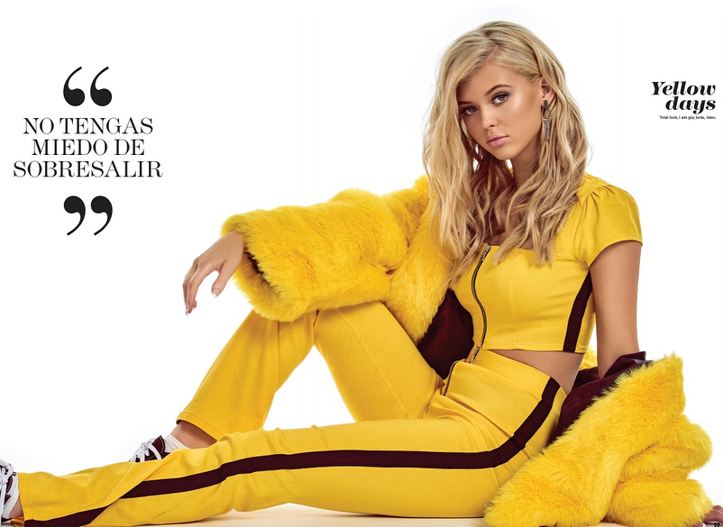 a young woman with blonde hair and stylish earrings is sitting crosslegged on a white surface in a fashion photoshoot. she is wearing a vibrant yellow outfit consisting of a shortsleeved crop top with a zipper and matching track pants with a dark brown stripe down the side. over the outfit, she has draped a fluffy yellow fur jacket, adding a touch of luxury and texture. a quote in spanish appears on the left side of the image: "no tengas miedo de sobresalir," which translates to "don't be afraid to stand out". the words "yellow days" appear in the top right corner of the image.