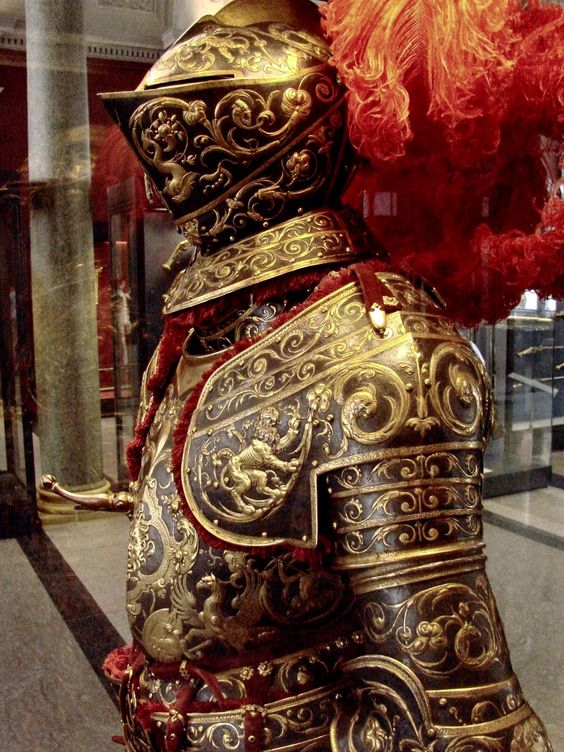 a closeup view of an ornate suit of armor displayed on a mannequin in a museum. the armor is predominantly gold with intricate etched designs and sculpted figures. a bright red plume adorns the helmet, adding a splash of color. the lighting is artificial, likely from spotlights within the museum setting.  the perspective is eyelevel, giving the viewer a sense of the impressive craftsmanship and detail of the armor.