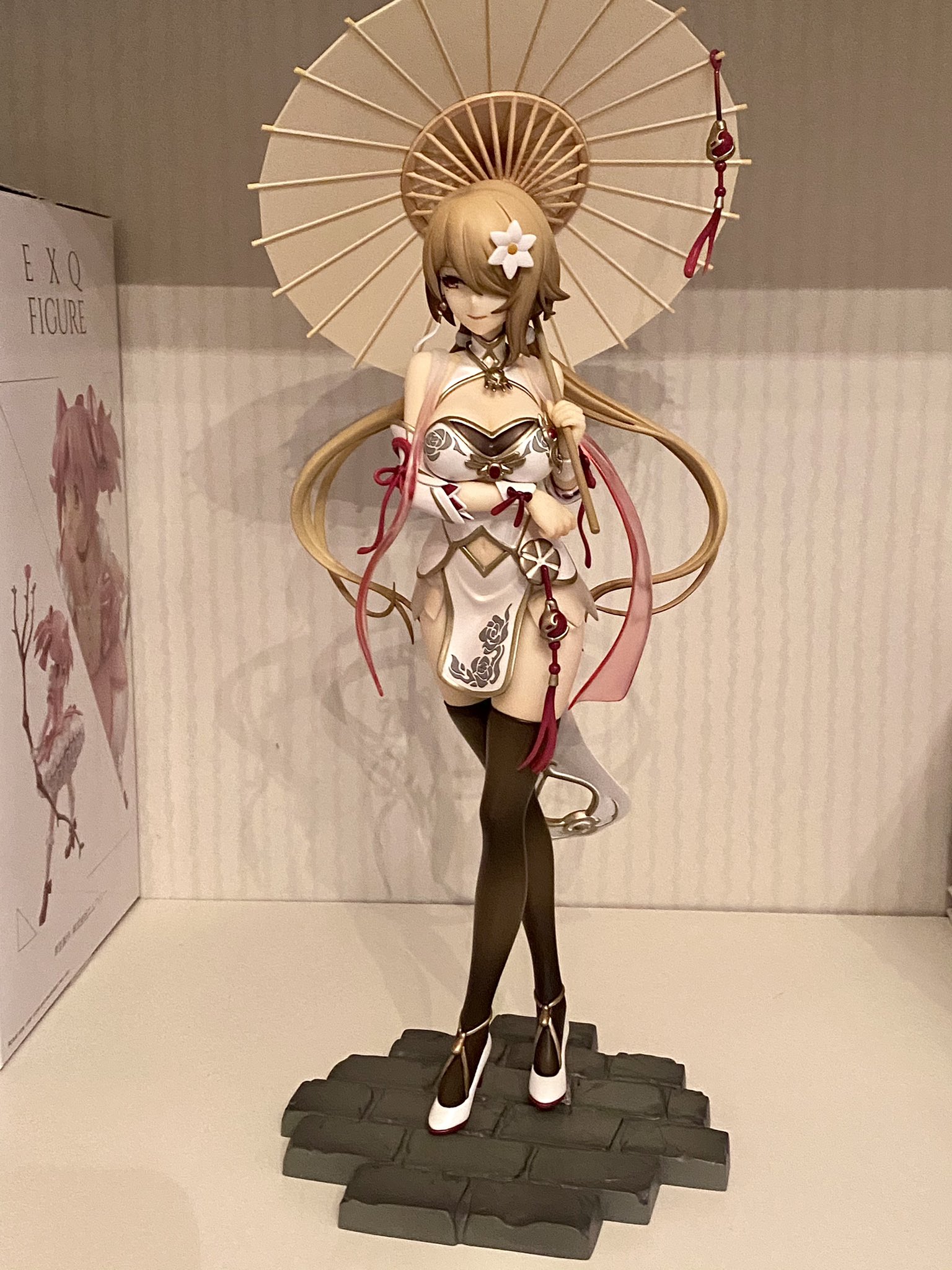a closeup of a beautifully crafted anime figurine of a young woman in a white kimono with pink and red accents. she is holding a parasol and smiling softly. the figurine is standing on a stone base and is surrounded by a white wall. the background is a white wall.