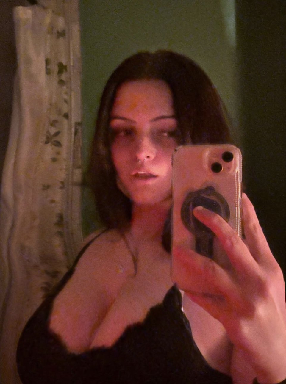 closeup selfie of a young woman with long dark hair. she's wearing minimal makeup and a black lace bralette.  the lighting is dim and warm, creating a moody atmosphere.  she's holding a phone to take the picture. the overall aesthetic is sensual and slightly mysterious.