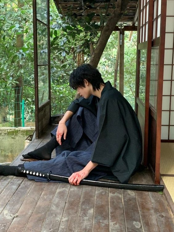 a young man dressed in a traditional japanese hakama and kimono sits dejectedly on a wooden porch, his hand resting on a katana sword laid out in front of him. the setting appears to be a traditional japanese exterior, with lush greenery visible through the open porch. the lighting is natural, casting soft shadows across the scene.