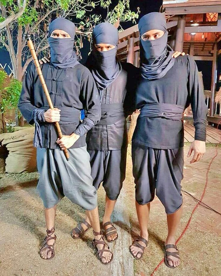 the image shows three men, all wearing black clothes. they are standing together, their faces are covered with a black cloth. they are holding a wooden stick and are looking at the camera. they are standing outdoors, and they are wearing brown sandals.