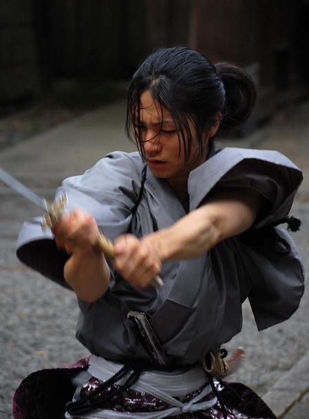 a woman, dressed in traditional japanese clothing resembling that of a samurai warrior, is captured midswing, brandishing a katana sword. her face is contorted with intense focus and determination as she grips the hilt with both hands. the setting appears to be an outdoor street, likely an attempt to recreate a historical japanese setting. the overcast lighting casts a dramatic shadow on the ground and emphasizes the intensity of the moment.