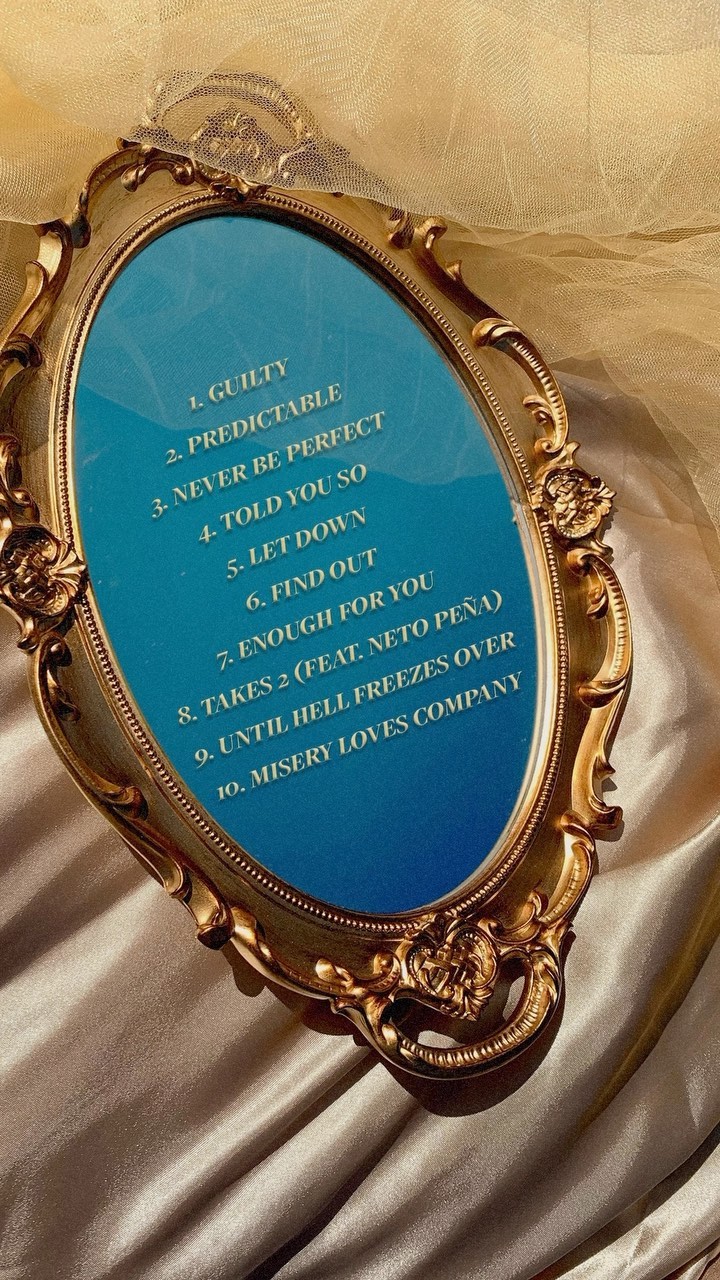 an ovalshaped mirror with an ornate gold frame displays a tracklist against a blue background. the list includes song titles such as "guilty," "predictable," and "takes 2 (feat. neto peña)." the mirror rests on a surface covered with draped satin fabric.