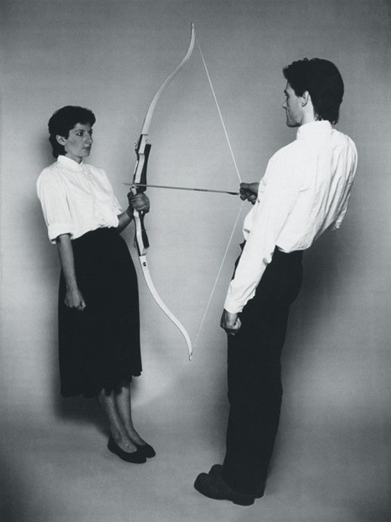 a black and white photograph depicting a performance art piece. a man and a woman are positioned facing each other, sharing a bow and arrow. the woman holds the bow, while the man seems to be guiding her aim. both wear simple, formal attire  the woman in a white shirt and black skirt, the man in a white shirt and black pants. the setting appears to be a minimalist studio. the overall mood is one of tension and implied interaction, possibly exploring themes of relationships, power dynamics, or control.  the style is reminiscent of 1980s performance and conceptual art.