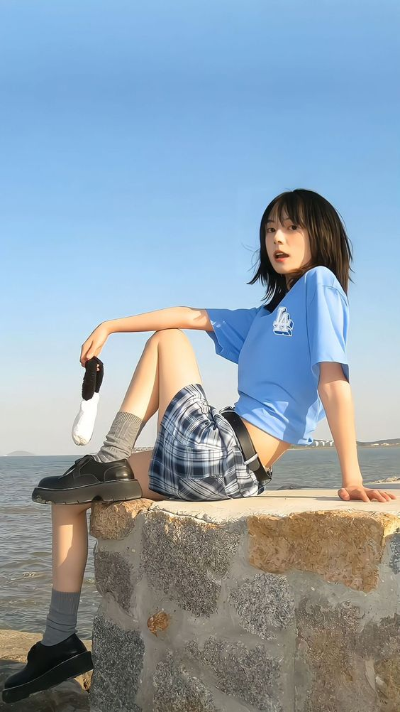 a young woman with short black hair is sitting on a rock wall facing towards the camera, with her body slightly angled to the left. she is wearing a blue tshirt, a blue and white plaid skirt, black socks, and black shoes. she is holding a white sock in her right hand. the background is a clear blue sky and a calm ocean.