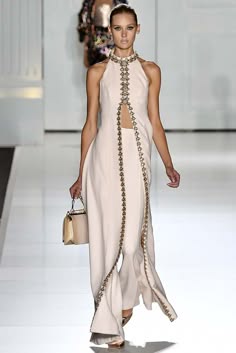 a fashion model walks the runway in a long, elegant white dress with intricate detailing. the dress is sleeveless and has a high neckline, and the skirt is flowing and dramatic. the model is wearing high heels and has her hair pulled back from her face. she looks confident and sophisticated as she poses for the cameras.