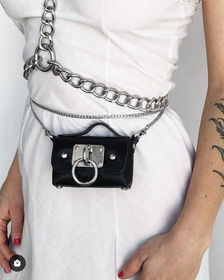 a woman wearing a white dress with a black belt bag that has a silver ring detail and is attached to a silver chain around her waist. she has a tattoo on her arm and red nail polish. the image is a closeup shot of the outfit.