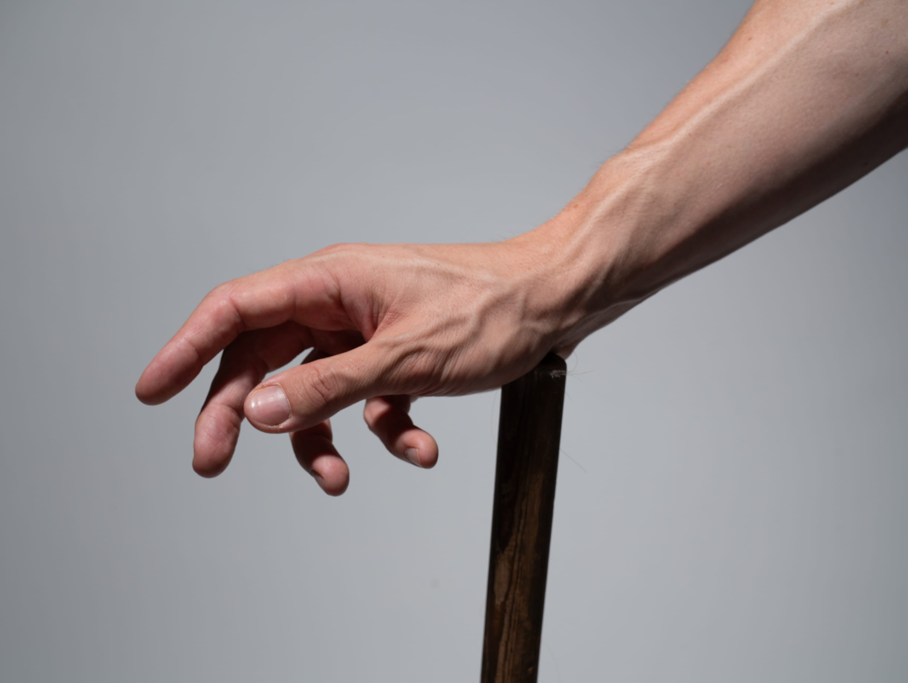 a closeup side view of a man's hand tightly gripping a wooden pole against a plain, light grey background. the studio lighting highlights the muscular structure and prominent veins of the arm, emphasizing strength and tension in the grip.