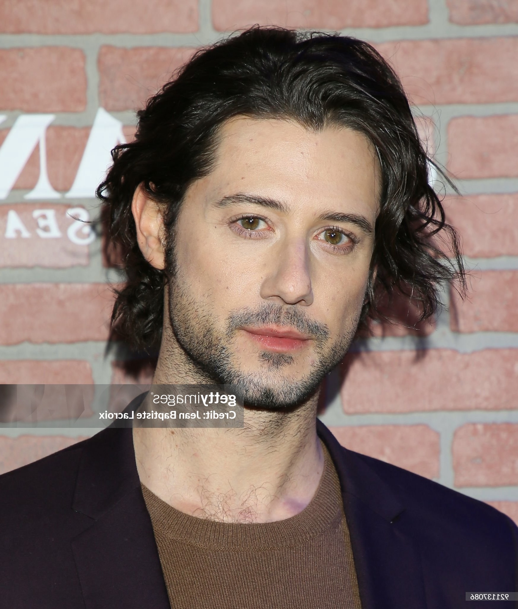 this is a headshot of a man with dark hair and a beard. he is wearing a brown shirt and a black jacket. he is standing in front of a brick wall with a sign on it. the lighting is soft and the perspective is eye level.