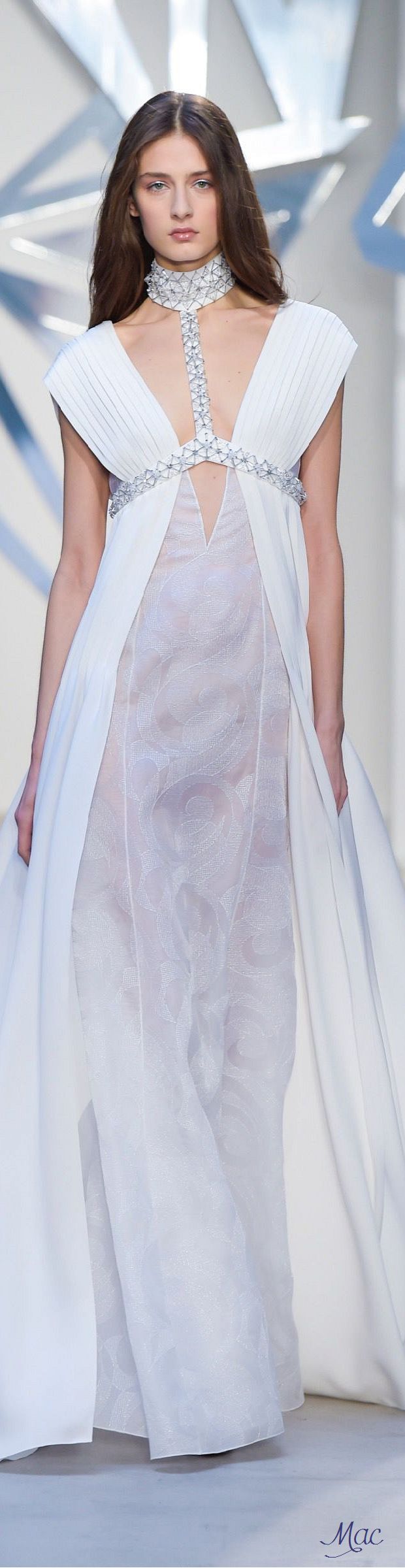 a beautiful model with long brown hair is wearing a long white gown with a  necklace and posing elegantly on a runway. the gown has a flowing skirt and a sheer bodice. she is looking directly at the camera with a confident and sophisticated expression. the background is blurry, but the focus is on the model and her outfit. the lighting is soft and flattering, creating a sense of luxury and glamour.