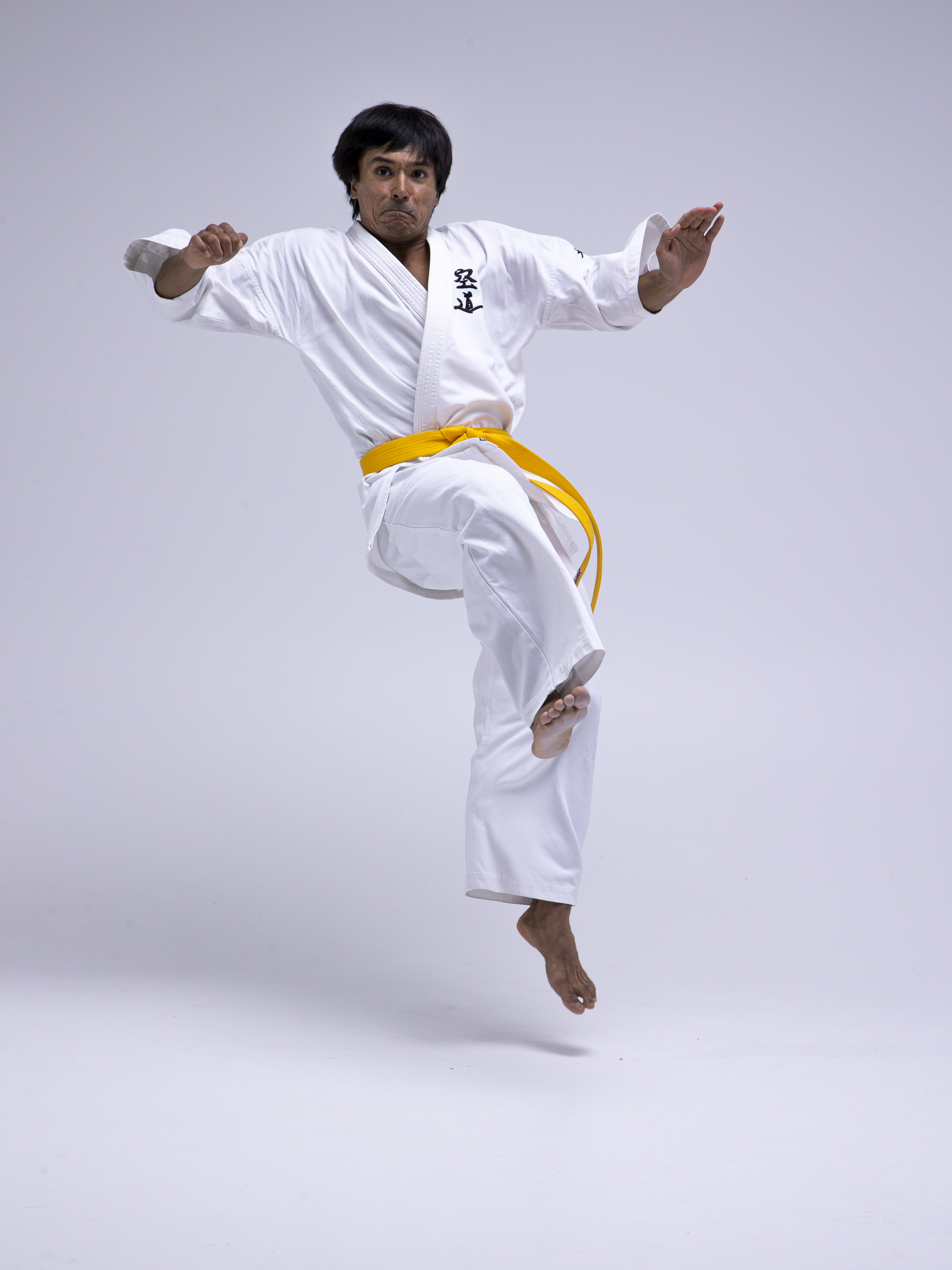 a man in a white karate uniform with a yellow belt is captured midair in a dynamic jumping pose. he is barefoot and stands against a plain white background.