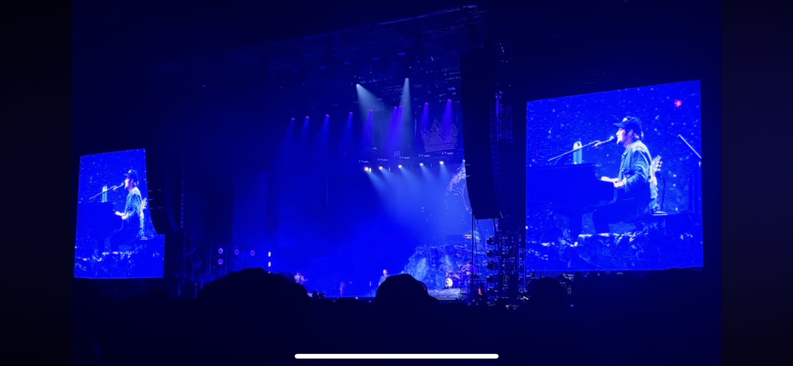 this image shows a concert stage with two screens displaying a musician at a piano, in the background. the stage is dark blue with spotlights, the crowd is a silhouette in the foreground, and the screen displays the performer in the center.