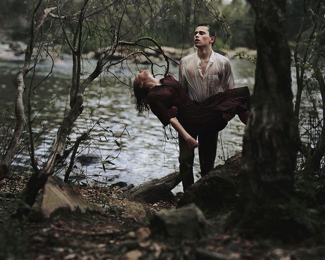 in a dimly lit forest, a young man wades through the shallows of a river, carrying a limp woman in his arms. the scene is heavy with emotion, suggesting a narrative of love, loss, and despair. the man's expression is pained yet determined, while the woman's lifeless posture suggests a tragic outcome. the natural environment, rendered in muted greens and browns, enhances the somber and evocative mood of the image.