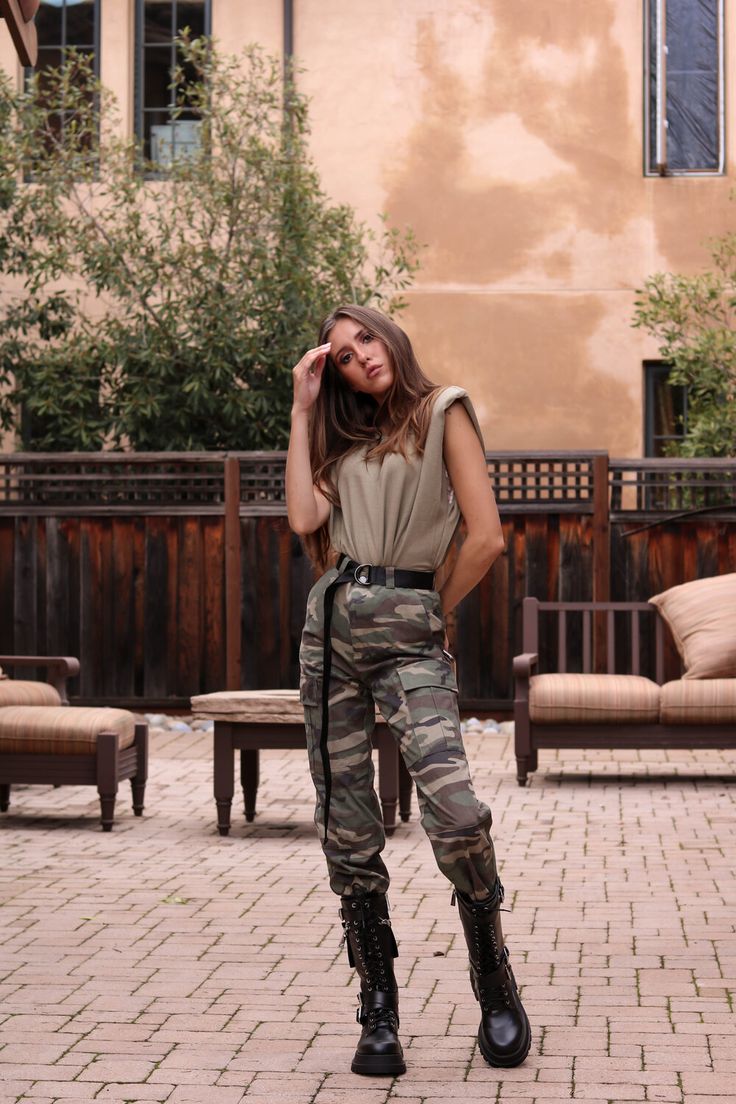 a young woman is posing outdoors in a courtyard setting. she's wearing a stylish olive green sleeveless top paired with camouflage cargo pants and black combat boots. she has a relaxed yet confident demeanor, one hand gently touching her hair.  the background features a brick patio, wooden furniture, and a tancolored building with trees. the overall aesthetic is casual, fashionable, and slightly autumnal.