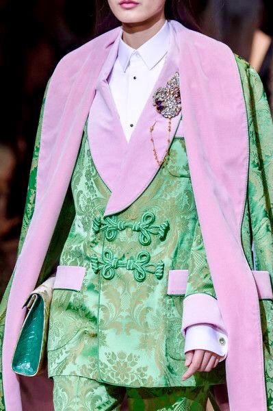 a woman wearing a light green brocade jacket with pink lapels and a white blouse underneath. the jacket features floral patterns and oriental knot closures. a brooch is pinned to the lapel. the woman is wearing a light green satin jacket with light green floral brocade all over and a light pink shawl.