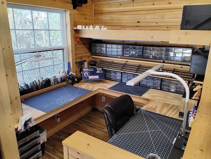 an organized craft room or hobby workspace with wooden paneling. the room features a ushaped desk with multiple cutting mats, storage bins above the desk, a window providing natural light, a desk lamp, an office chair, a desk organizer, and a giraffe figurine.