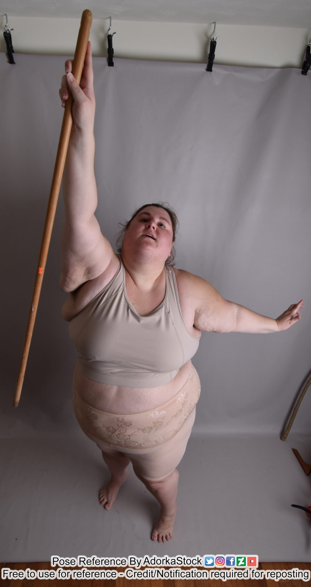 a woman in a sports bra and shorts holds a wooden staff above her head, one leg bent at the knee, the other leg straight, her upper body facing to the left. her arms are raised, holding the staff at the top.  the background is a light grey, with the hooks and ceiling visible in the top left corner.