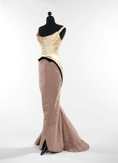 a full shot of a formal gown on a black mannequin against a white background. the dress has a strapless beige satin bodice with black trim at the waist. the skirt is a light brown color and fitted until the knees where it flares out. there is a slit at the bottom of the left side of the skirt.
