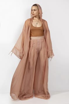 a woman is posing in a long pink dress with long sleeves against a white background. she is looking forward with her hands at her sides. the dress is loosefitting and flowy. the woman is standing with her weight on her left leg.