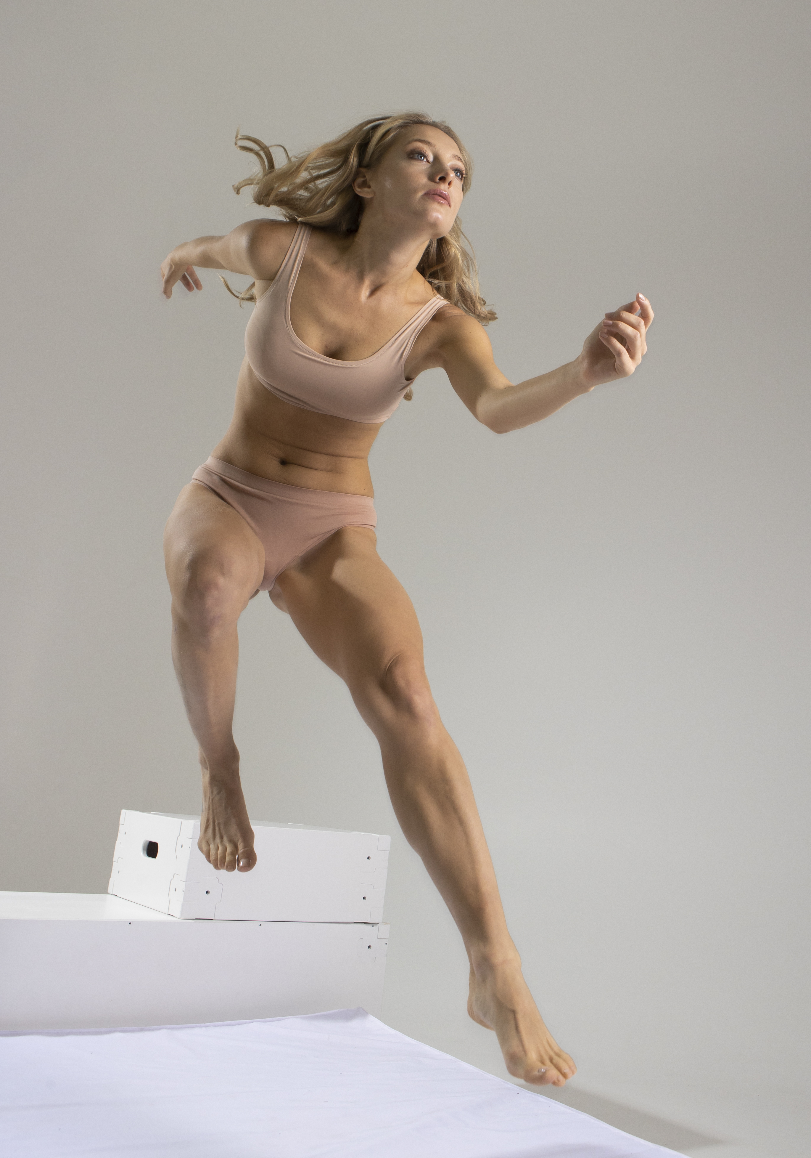 a full shot depicts a woman in a dynamic pose, captured midjump while descending a set of white stairs. she is wearing matching underwear and a sports bra in a nude tone. the woman is blonde and her hair is flying behind her. her physique is athletic, revealing toned muscles and a strong build. she is barefoot, further emphasizing the active and free movement of the scene. the backdrop is a plain studio setting, ensuring the focus remains on the woman and her action.