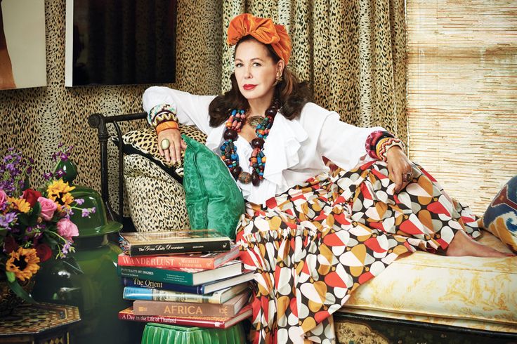 a stylish woman in her 50s or 60s, adorned in an orange turban and a colorful, patterned skirt, lounges on a chaise lounge. the room is elegantly decorated in a bohemian style with rich colors, patterned textiles, and leopard print wallpaper. the woman is wearing numerous bracelets and a long necklace. she looks relaxed and content surrounded by luxury and various home decor items, including a stack of books and a bouquet of flowers. the overall aesthetic is one of eclectic, highend design and relaxed sophistication.