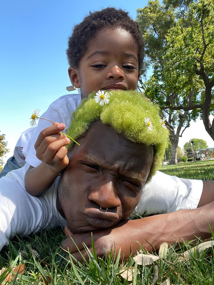 a young black man with green hair is laying down on the grass in a park, with a young black boy on top of him smiling. the boy is holding a daisy and there are flowers in the man's hair. they both appear to be very happy and enjoying each other's company.