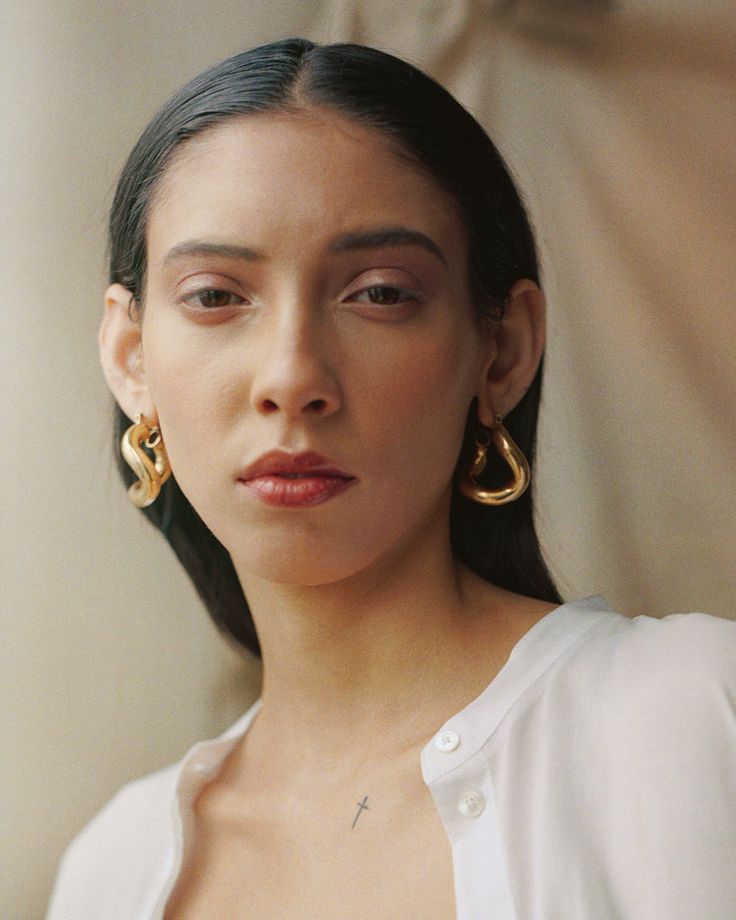 a portrait of a woman with dark hair, wearing gold earrings and a white shirt. she has a small cross tattoo on her chest. the background is a neutral color.